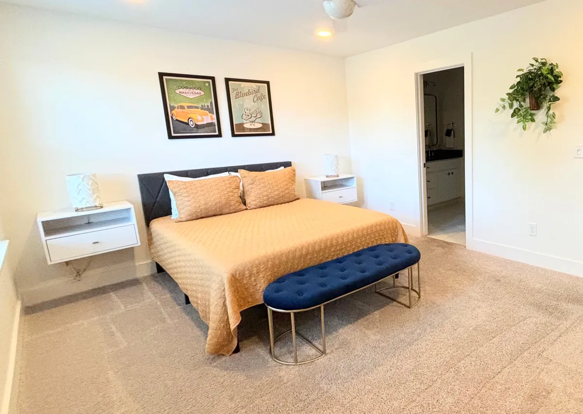 A queen bedroom with a decorative quilt on the bed, a blue upholstered bench at the foot, and artwork adorning the walls. The room is illuminated by natural light, creating a serene and cozy atmosphere ideal for rest and relaxation.