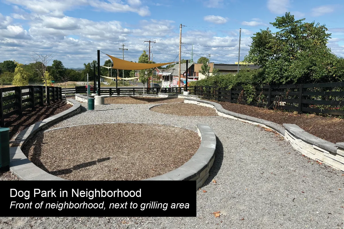 An inviting outdoor dog park enclosed by fencing, with gravel pathways, a shade structure, and adjacent grilling area. The space is designed for socializing, dog play, and family gatherings.