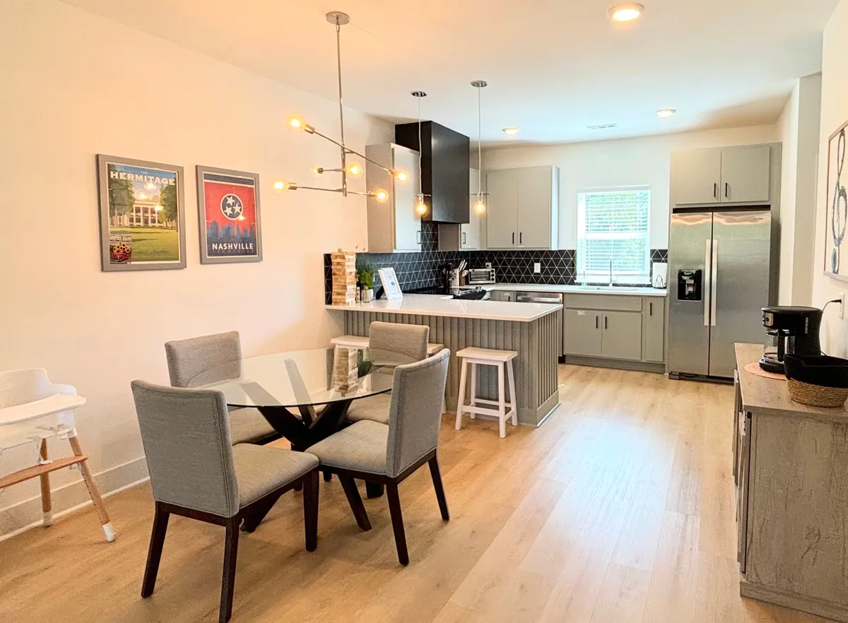 A modern, welcoming dining space with a glass-topped table, contemporary light fixture, kitchen cabinetry visible in the background, and decorative art, emphasizing both style and function.