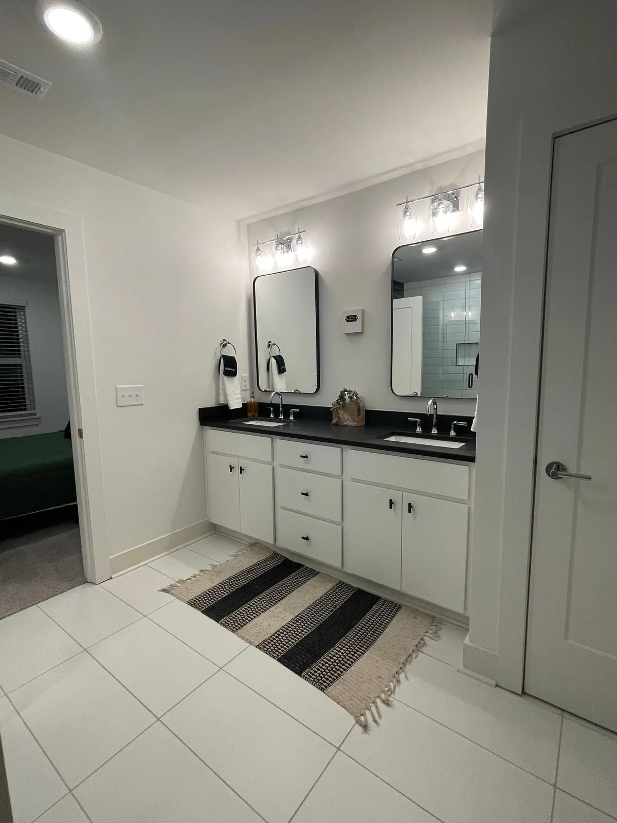 A sophisticated bathroom scene featuring dual sinks, mirrors, a black countertop, and a decorative rug. The layout suggests plenty of space for personal grooming and self-care routines, supporting the narrative of convenience and luxury.