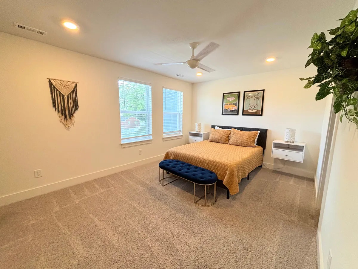 A warmly lit queen bedroom featuring a bed with a textured golden coverlet, a blue tufted ottoman, framed wall art, hanging macramé decor, and large windows letting in ample natural light. The space feels tranquil and inviting, supporting relaxation and sleep.