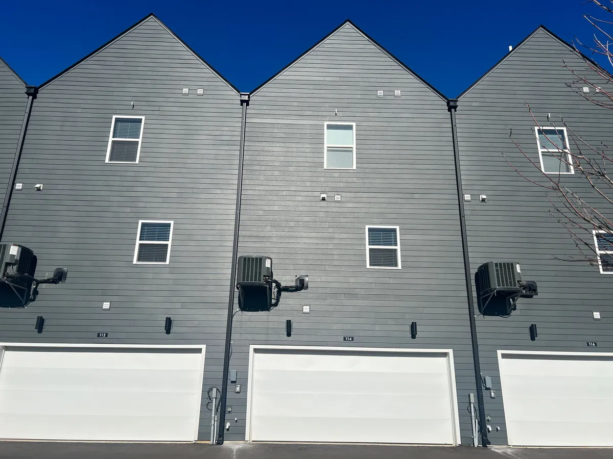 A contemporary exterior view of the property, featuring gray siding, white garage doors, and visible air conditioning units. The scene suggests a well-maintained and safe neighborhood, ideal for both arrivals and departures, and gives a sense of the surrounding community environment.