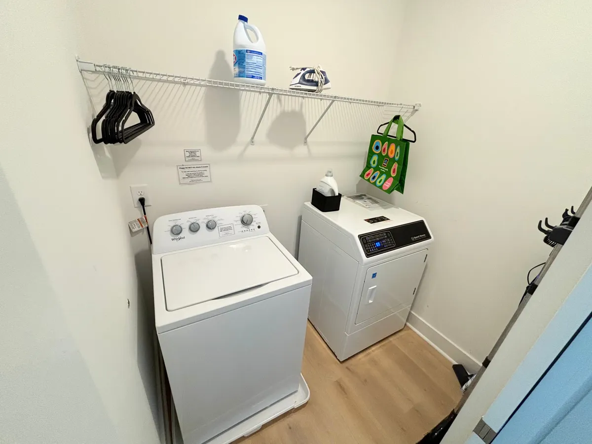 A dedicated laundry area featuring a front-loading washing machine and dryer, shelf with hangers, and cleaning supplies, all arranged in a compact, well-organized space.